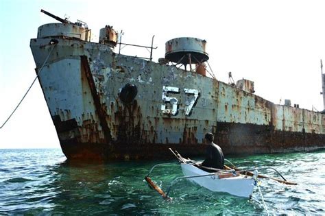 a man in a rowboat paddling past an old rusted ship on the ocean