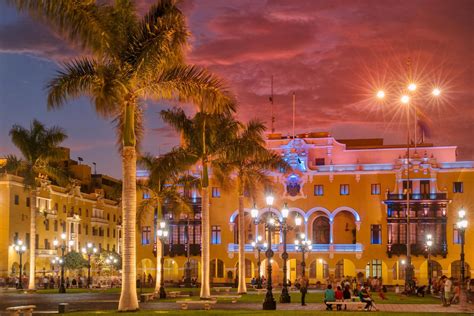 people are sitting on benches in front of a building at night with palm trees and street lights