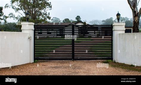 Black modern metal driveway property entrance gates set in concrete fence, lights, trees Stock ...