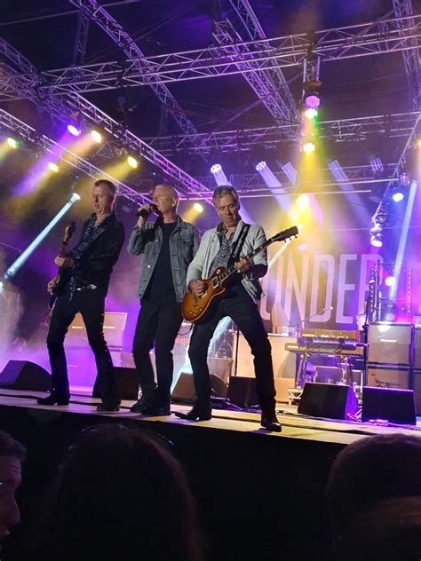 three men standing on stage with guitars in front of purple and green lights, one man is singing ...