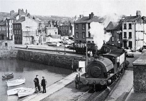 black and white photograph of steam engine train on tracks next to water with small boats in ...