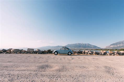 vintage blue volkswagen beetle parked on road in front of lake with mountains in the distance ...