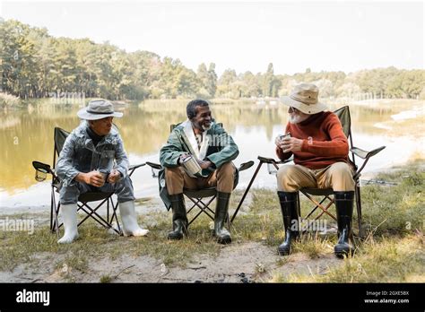 Cheerful multicultural men in fishing outfit holding thermo cups near ...