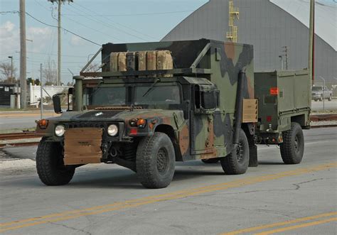 an army truck driving down the road with another vehicle behind it in front of a building