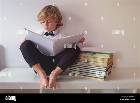 Full body of concentrated barefoot boy in formal wear with bow tie reading book while sitting on ...