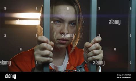 Female prisoner in orange uniform stands in prison cell. Scared woman criminal shakes, holds ...