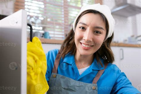 Smiling young woman in apron and cleaning gloves open kitchen cabinets ...