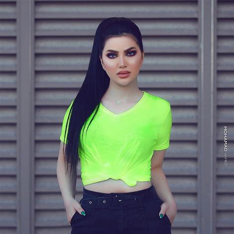 a woman with long black hair wearing a neon green crop top and jeans, standing in front of a ...