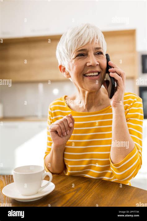 Cheerful mature woman talking on mobile phone and drinking coffee at home at dining table Stock ...