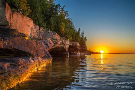 the sun is setting behind some rocks on the water's edge, with trees in ...