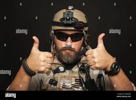 Brutal man in military desert uniform and body armor shows two fingers up on black background in ...