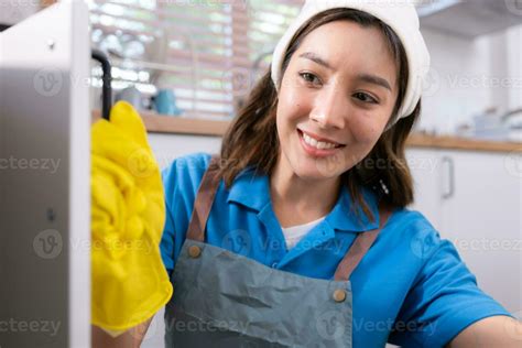 Smiling young woman in apron and cleaning gloves open kitchen cabinets ...