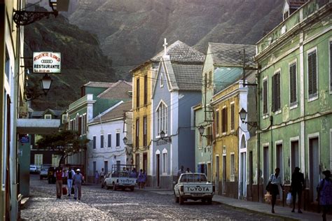 an old car parked on the side of a road in front of buildings with mountains in the background