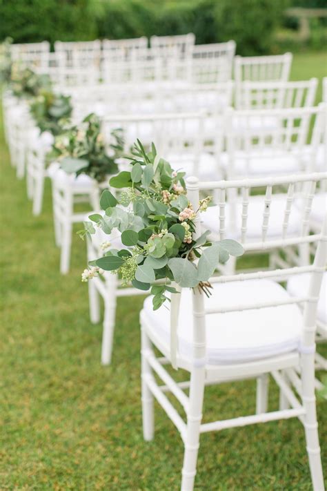 Elegantly lined white wedding ceremony chairs accented by Rustic Italian greenery to line the ai ...