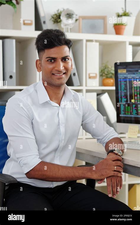 Portrait of handsome young Indian trader in white shirt sitting at office table with stock ...