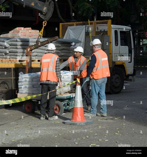 Men unloading palletised bagged building materials off delivery truck adjacent building site ...