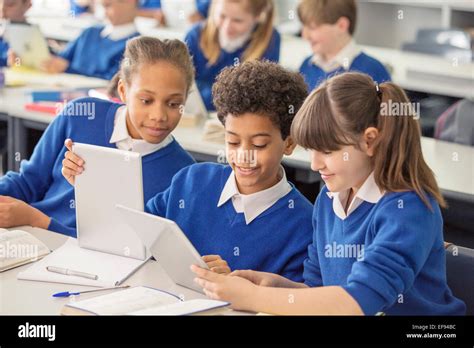 Elementary school children wearing blue school uniforms using digital tablets at desk in ...