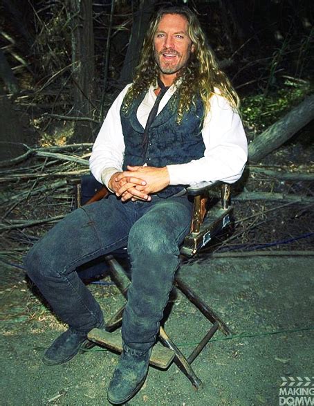 a man with long hair sitting on a chair in the woods wearing a vest and jeans