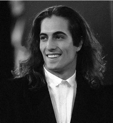 a man with long hair wearing a black suit and white shirt smiles at the camera