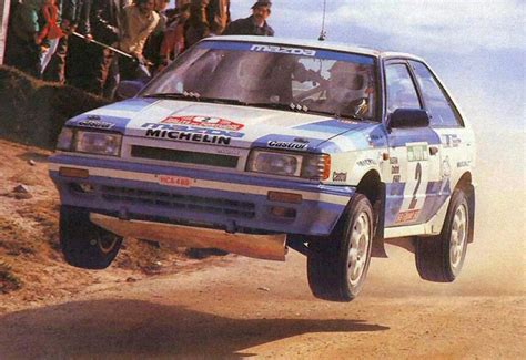 a blue and white rally car making a jump on dirt road with people watching from the side