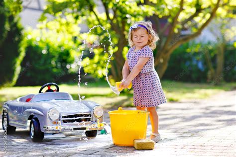 Cute gorgeous toddler girl washing big old toy car in summer garden, outdoors. Happy healthy ...