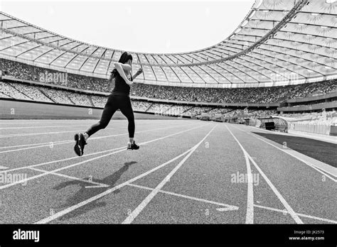 Back view of young fitness woman in sportswear sprinting on running track stadium, black and ...