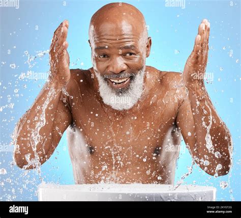 Water splash, senior and black man cleaning face in studio isolated on a blue background ...