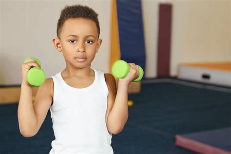 Cute boy athlete of African appearance doing physical exercises at gym after school, holding two ...