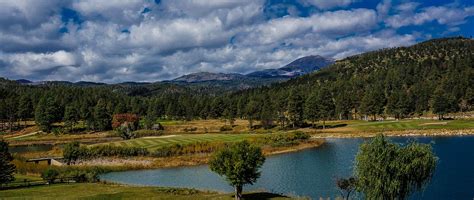 a lake surrounded by trees and mountains under a cloudy blue sky with white fluffy clouds