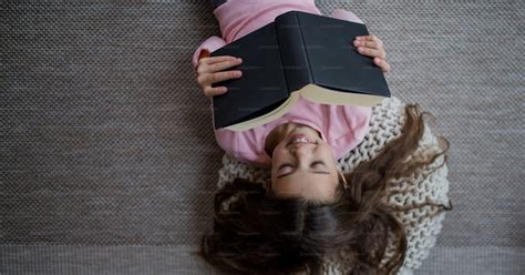 A top view of happy little girl lying on floor and reading book indoors ...