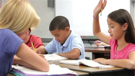 Female Student Working At Desk Raises Hand To Ask Teacher Question - Teacher Stand By Desk And ...