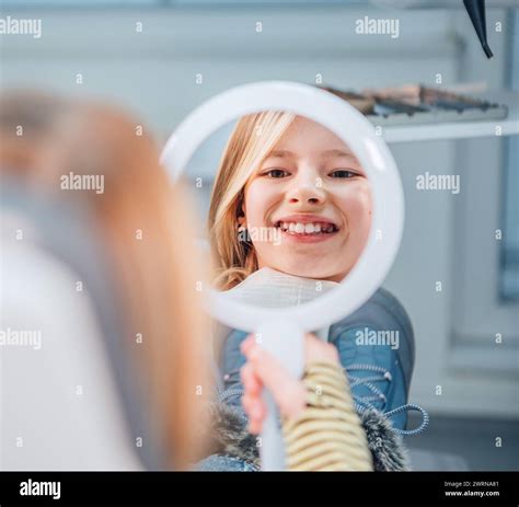 Little girl sitting in stomatology clinic chair and smiling at mirror showing her teeth after ...