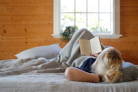 Image of Woman lying in bed reading book with timber walls and window in background - Austockphoto