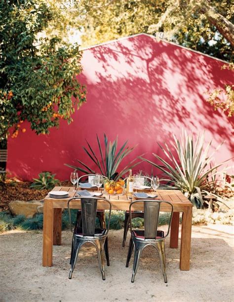 an outdoor dining table with chairs and potted plants in front of a red wall