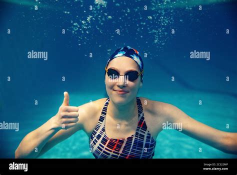 Female swimmer in swimsuit, swimming cap and glasses shows thumbs up underwater in pool. The ...