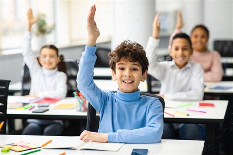 Diverse small schoolkids raising hands at classroom Stock Photo by Prostock-studio