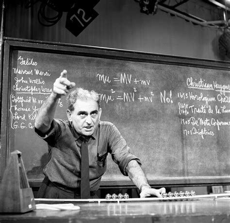 an old man in front of a blackboard with writing on it and pointing at something