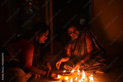 Two beautiful Indian Bengali women in Indian traditional dress are lighting Diwali diya/lamps ...