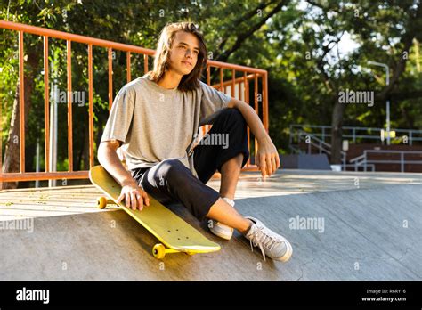 Photo of pleased skater boy 16-18 in casual wear sitting on ramp with skateboard in skate park ...