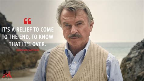 an older man wearing a sweater vest standing in front of some rocks on the beach