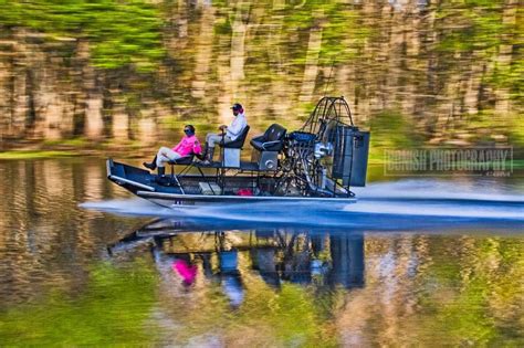 two people are riding on a boat in the water with trees behind them and one person is fishing