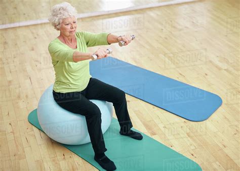 Senior female sitting on a fitness ball and lifting dumbbells. Old woman exercising with weights ...