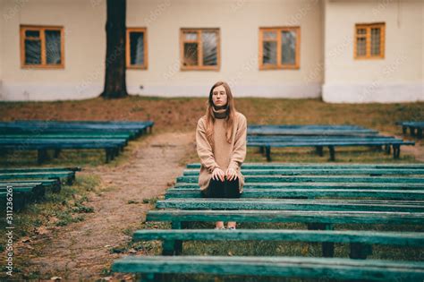 Sad depressed girl sitting alone on bench in park with multiple desolate benches around her ...