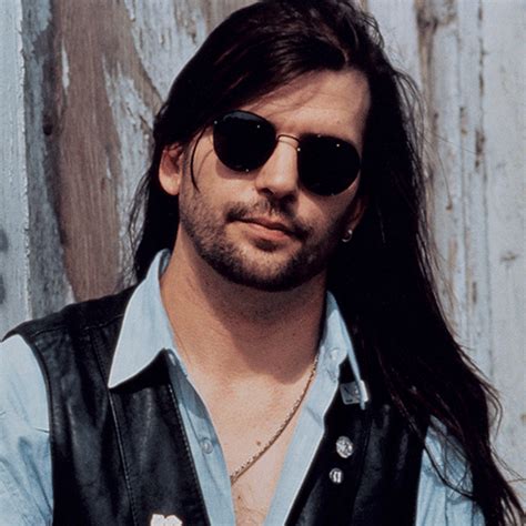 a man with long hair and sunglasses standing in front of a wooden wall wearing a leather vest