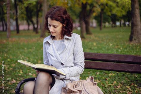 Side view of pleased brunette woman sitting on bench and reading book ...