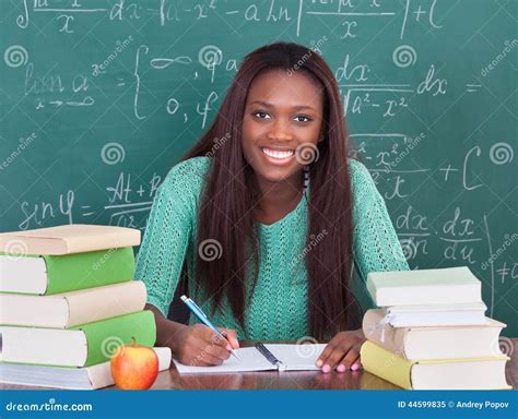 Confident Female Teacher Writing In Book At Classroom Desk Royalty-Free ...