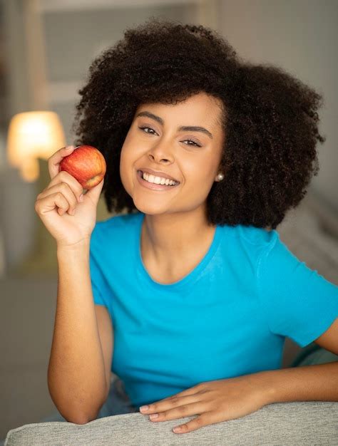 Premium Photo | Cheerful millennial african american curly lady enjoy apple on sofa in living ...
