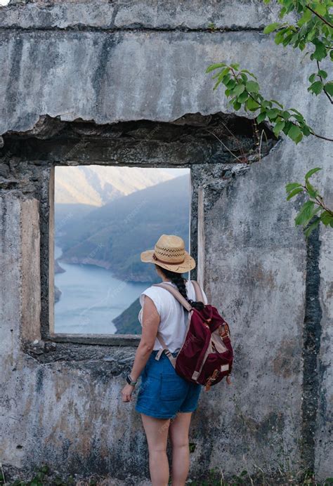 Premium Photo | Young girl with backpack and hat looks out of ruined window at mountainous ...