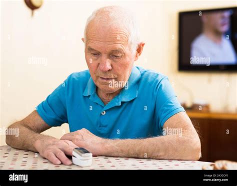 Focused worried elderly man measuring himself oxygen saturation while sitting at home table ...