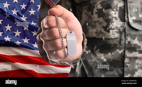 Male soldier holding military ID tag and American flag on background, closeup. Military service ...